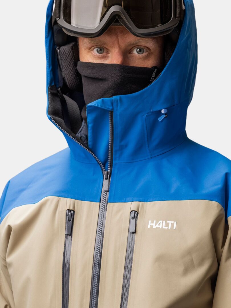 Henkilö, jolla on päällään hiihtokypärä, suojalasit ja musta Trone Fleece Tuubihuivi sinisen ja vaaleanruskean hiihtotakin alla.