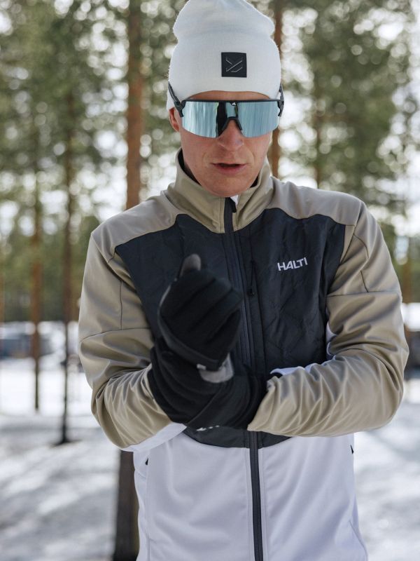 Talviurheiluvaatteisiin pukeutunut henkilö säätää Halti XC Touring II Hanskatiaan lumisessa metsässä.