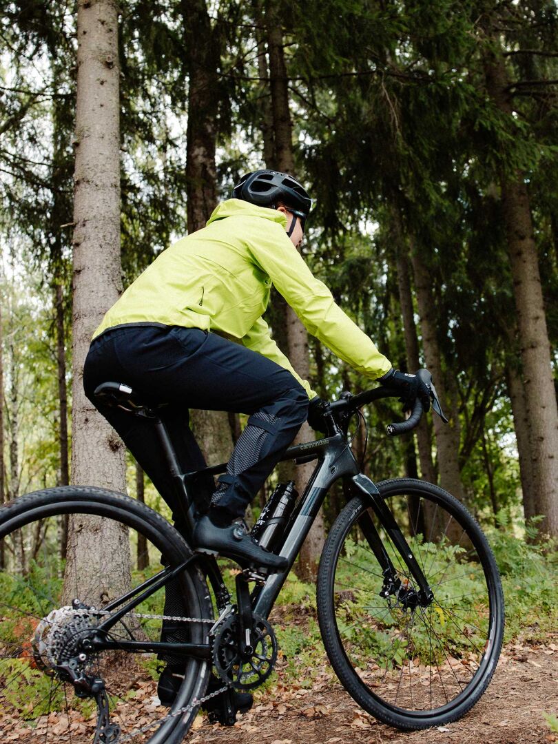Henkilö keltaisessa Halti Liike Naisten 3L DX Urheilutakki ja kypärä päällään pyöräilee metsän halki kulkevalla hiekkatiellä.