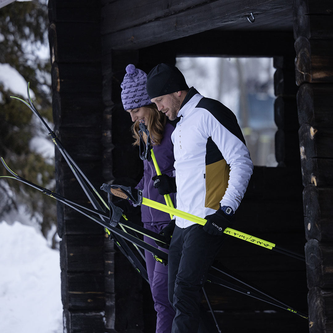 Kaksi ihmistä Halti Veloce Hiihtotakki Miesten -hiihtokeskuksessa pitelee suksia ja sauvoja puurakennuksen ulkopuolella lumessa.