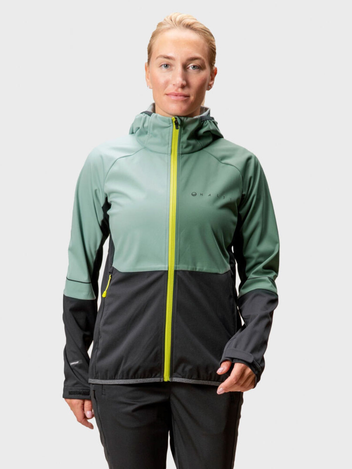 Henkilöllä on yllään Halti Exhale Stormwall Takki Naisten, vihreä ja musta softshell-takki, jossa on keltainen vetoketju.