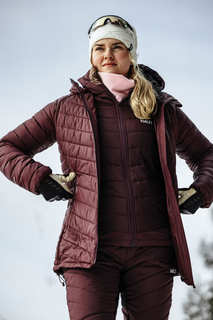 Henkilö, jolla on ruskeankeltainen Stormwall-takki ja Halti Hanki 2 Hybrid Hiihtohousut, seisoo ulkona talvivarusteissa.