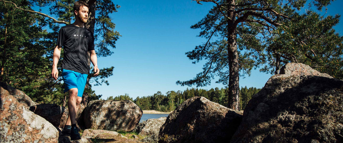 Mustaan paitaan ja sinisiin shortseihin pukeutunut mies kävelee suurten kivien keskellä metsäisellä alueella veden äärellä aurinkoisena päivänä.