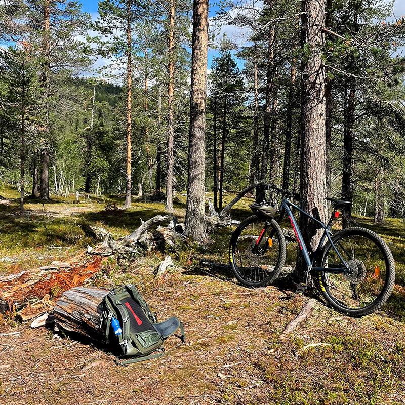 Polkupyörä nojaa metsän puuta vasten; reppu ja kypärä lepäävät maassa lähellä.