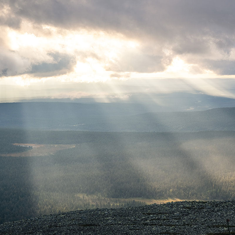 Auringonsäteet murtautuvat pilvien läpi metsäisessä maisemassa, jossa on kallioinen etuala ja kaukaisia kukkuloita.