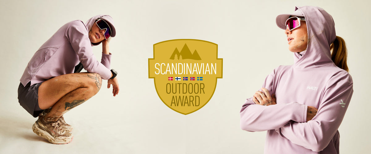 Laventelinväriseen huppariin, shortseihin ja aurinkolaseihin pukeutunut henkilö poseeraa Scandinavian Outdoor Award -logon vieressä valkoisella taustalla.