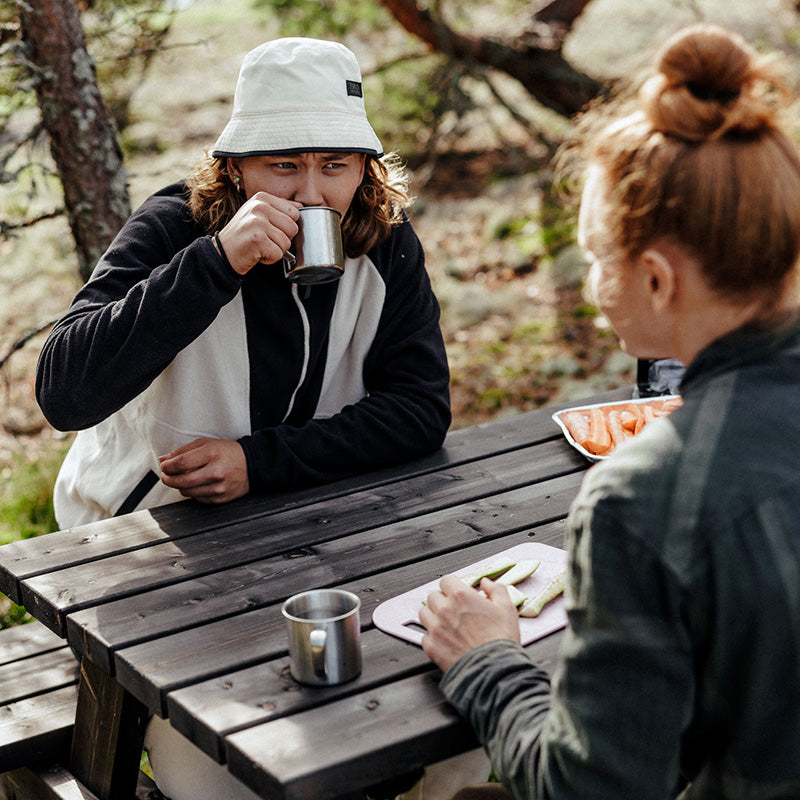 Kaksi ihmistä istuu ulkona piknik-pöydässä; toinen juo metallimaljasta, toinen syö viipaloitua ruokaa lautaselta.
